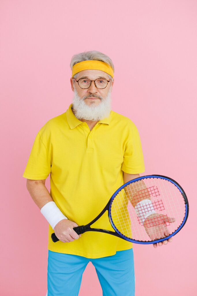 Elderly man holding a tennis racket against a vibrant pink background in a studio setting.