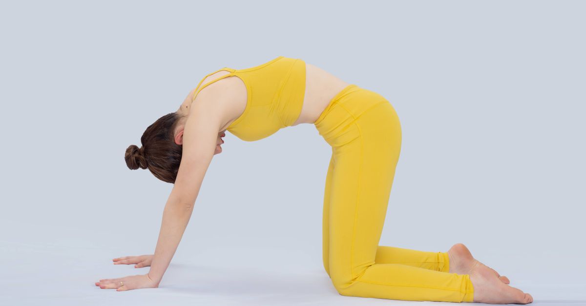 An adult woman in a yellow outfit performing cat pose yoga indoors, focusing on flexibility and well-being.