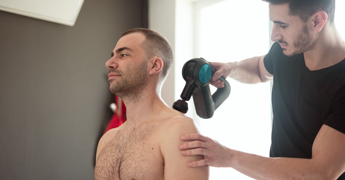 A physiotherapist provides shoulder massage therapy with a massage gun in a clinic.