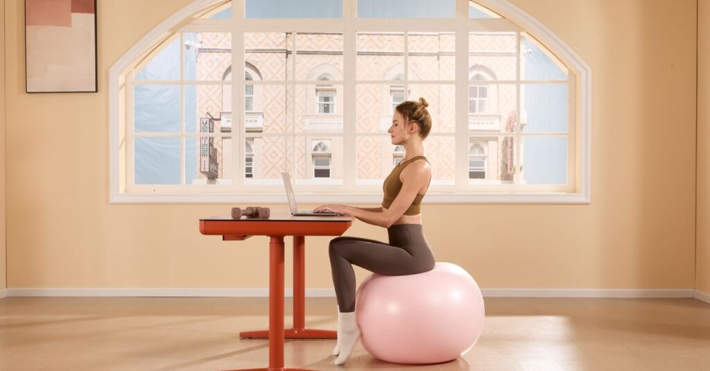 A young woman works on a laptop while sitting on an exercise ball in a bright, modern room.