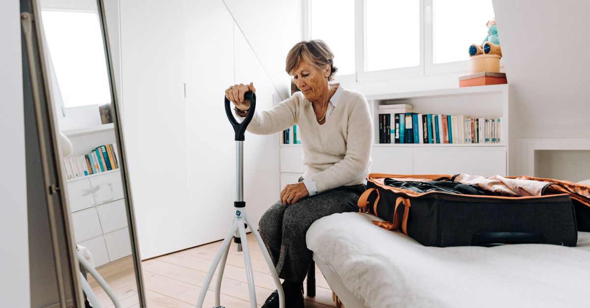 Senior woman sitting on bed with walker, preparing suitcase in bright bedroom.