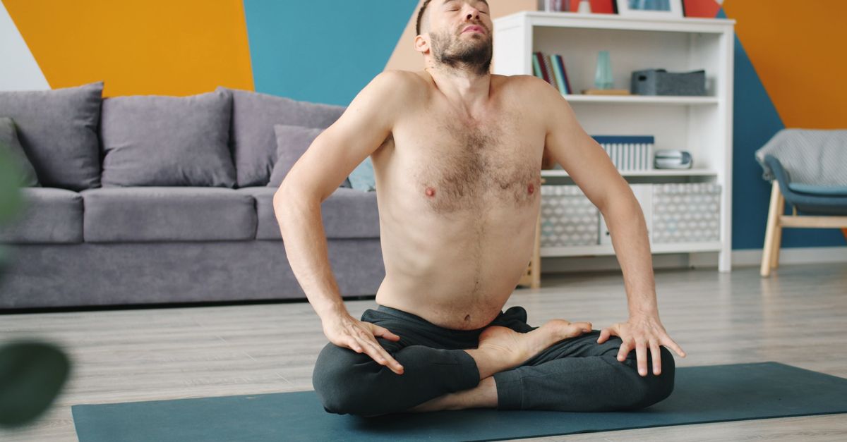 Shirtless man performing yoga meditation pose on mat in modern living room.