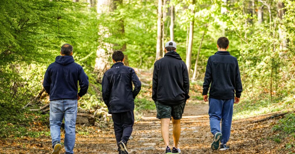 Four men in casual attire walking on a sunlit forest trail, enjoying nature.