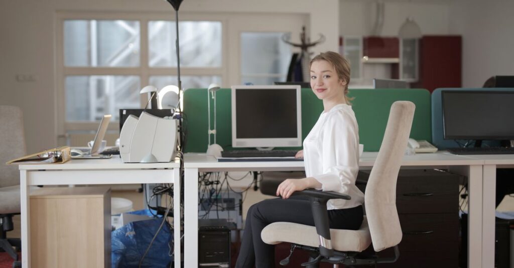 Side view of glad female worker in classy outfit sitting at table on office chair during work in modern workplace and looking at camera