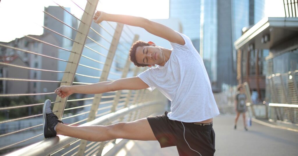 Calm focused African American male athlete with eyes closed in earphones and sportswear leaning on metal fence and doing side bend exercise while listening to music and stretching body on street against blurred urban environment in sunny day