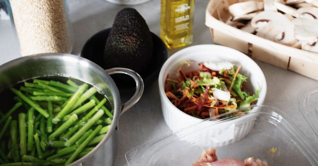 From above of raw chicken pieces in container green beans in pot of water and chopped champignons slices in wicker basket next to salad bowl avocado and spices in glass jar on countertop with various kitchen utensils
