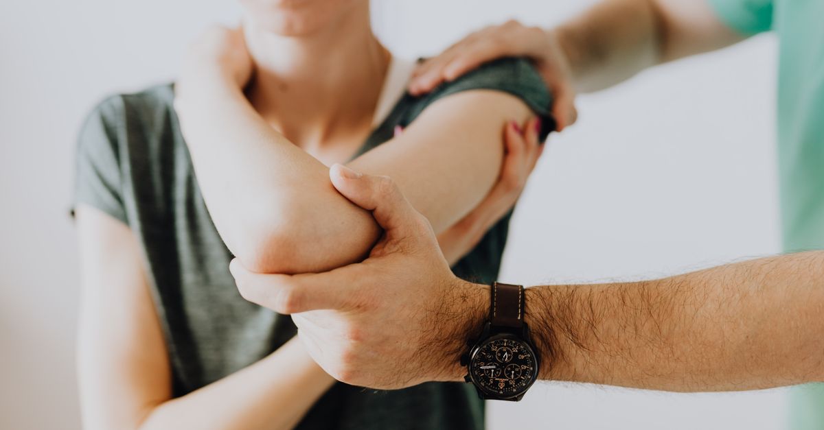 Crop faceless osteopath in modern wristwatch and uniform bending arm of anonymous thoughtful woman in casual wear in doctor office in hospital