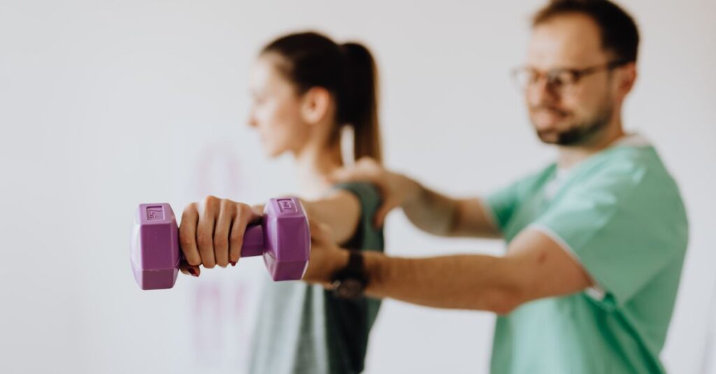 Side view of professional orthopedist in uniform and eyewear helping fit woman reaching arm with dumbbell in doctor office on blurred background