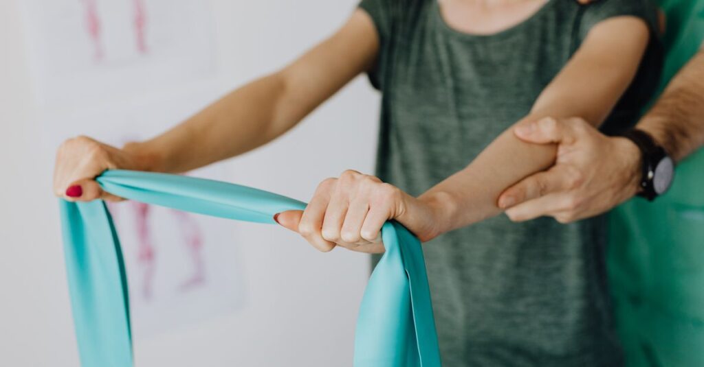 Crop unrecognizable orthopedist in uniform and wristwatch supporting shoulder and reached arm of faceless female patient with resistance band in doctor office