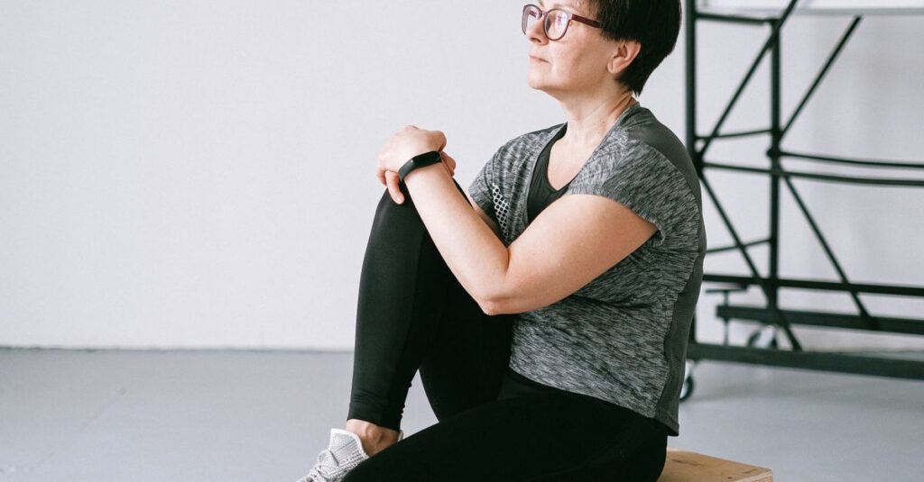 Senior woman in active wear sitting on a wooden box indoors, reflecting on wellness.