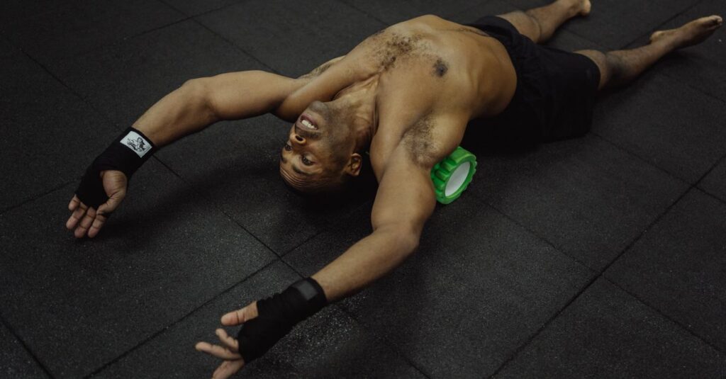 Topless man using foam roller for muscle recovery in gym setting.