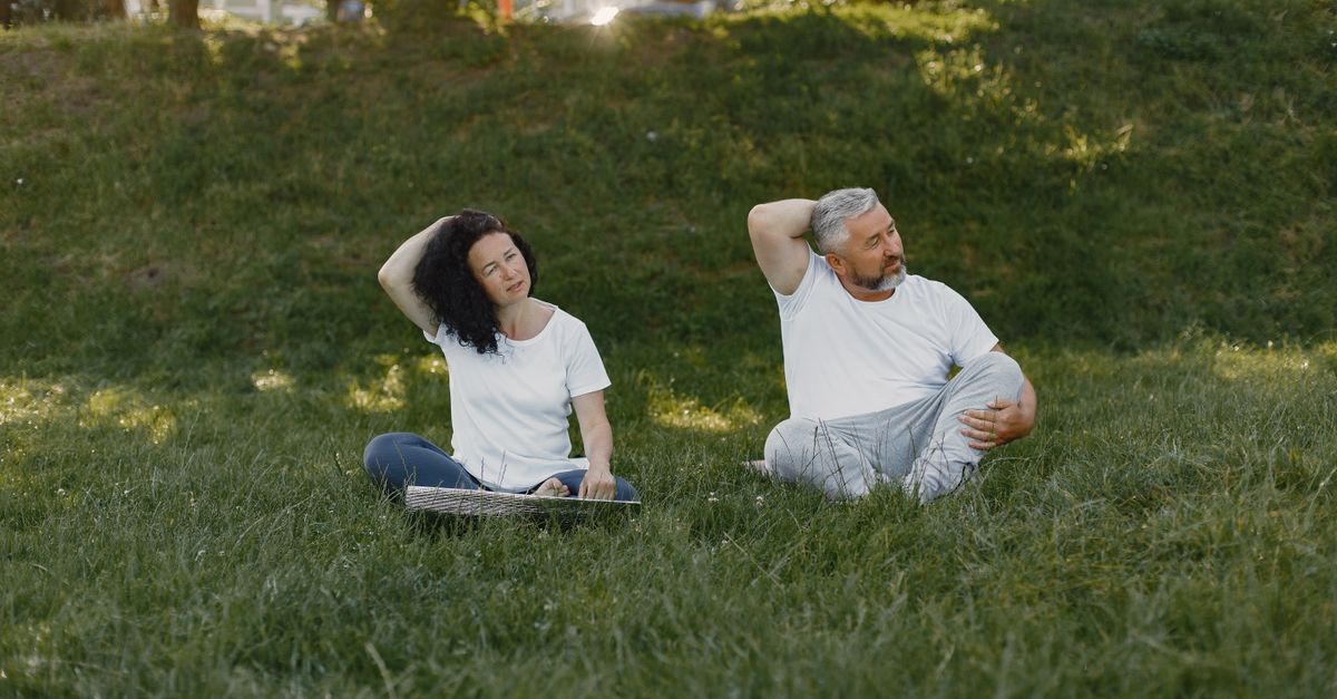 Couple doing neck stretches outdoors, promoting a healthy lifestyle in a sunny park setting.