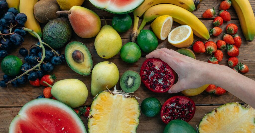 Colorful assortment of fresh fruits on wooden surface with a hand holding a pomegranate half.