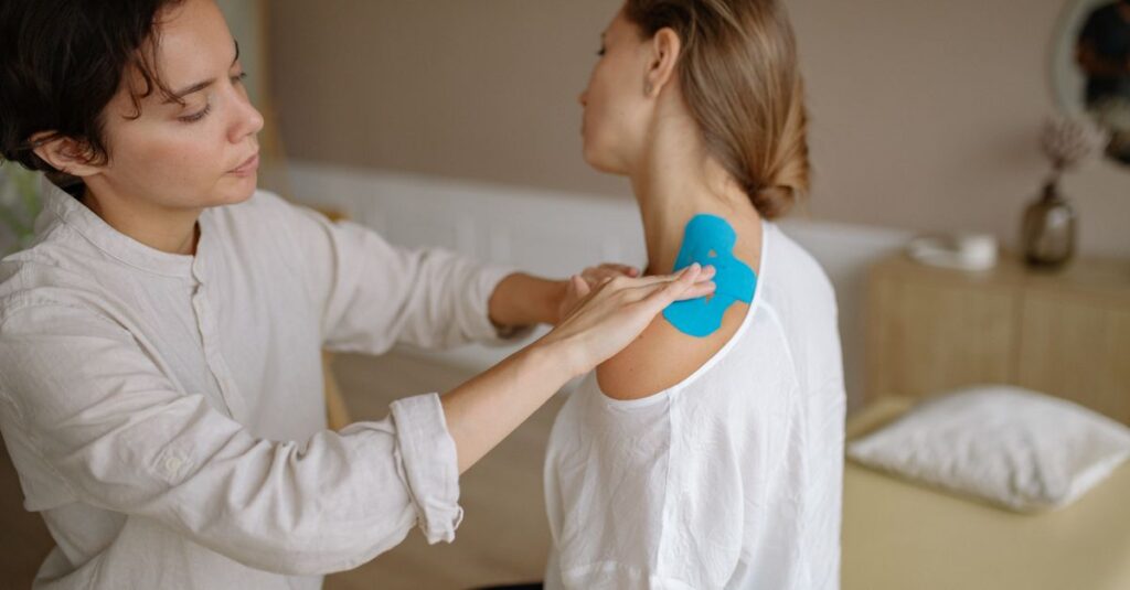 A physiotherapist applies blue kinesiology tape to a patient's shoulder for muscle support.