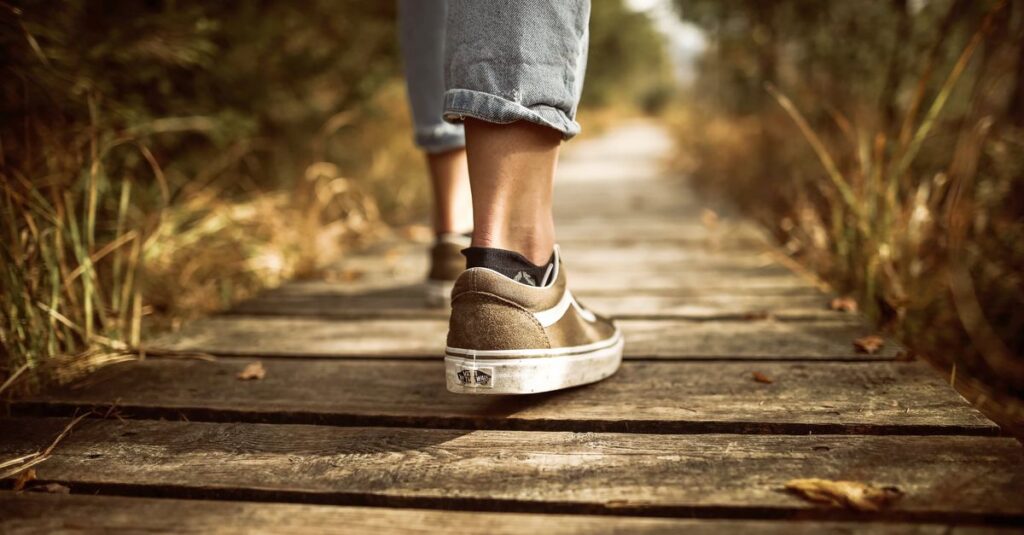 Person wearing sneakers walking on a wooden path in a sunlit park.