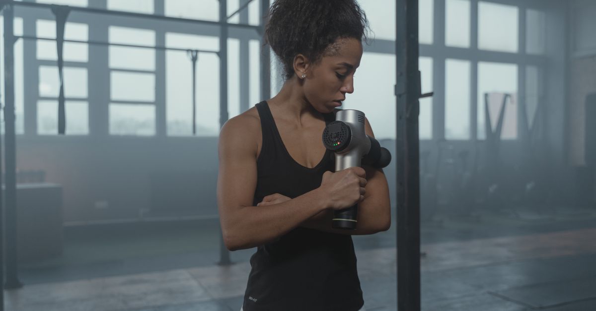Fit woman using a massage gun in a well-lit gym studio for muscle recovery.