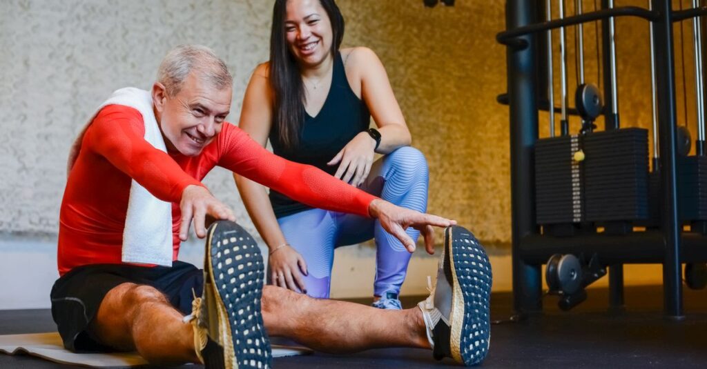 Elderly man in a gym stretching with a fitness coach, promoting a healthy lifestyle.