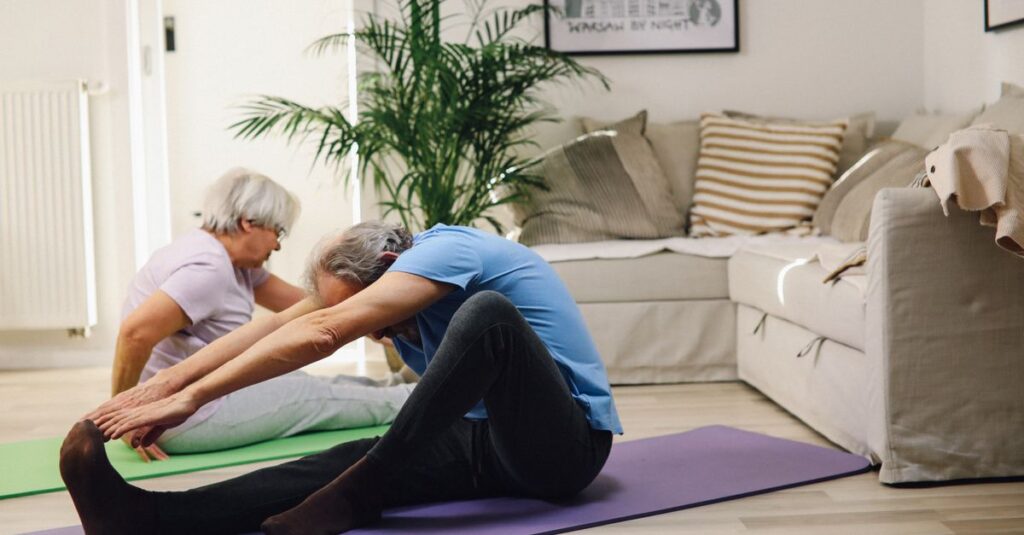 Two senior adults performing yoga stretches in a cozy living room setting.