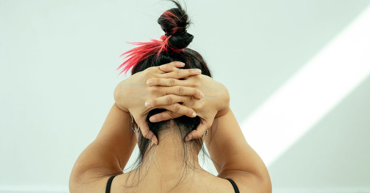 Back view of anonymous female stretching neck pressing clasped hands on head against white wall