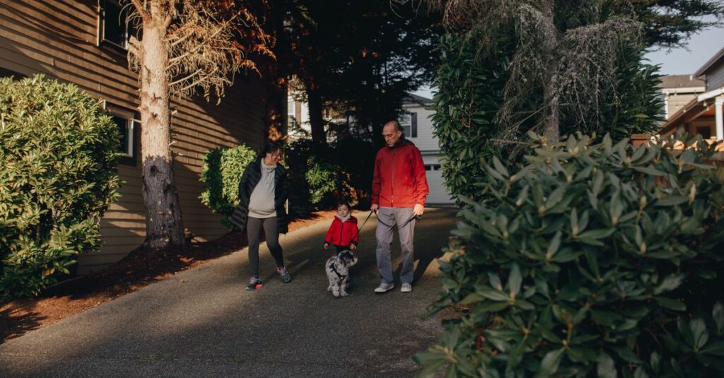 A family enjoys a walk with their pet dog on a sunny day, surrounded by lush greenery in a suburban neighborhood.