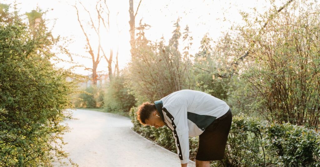 A man in sportswear bends over, stretching along a sunlit park path surrounded by trees.