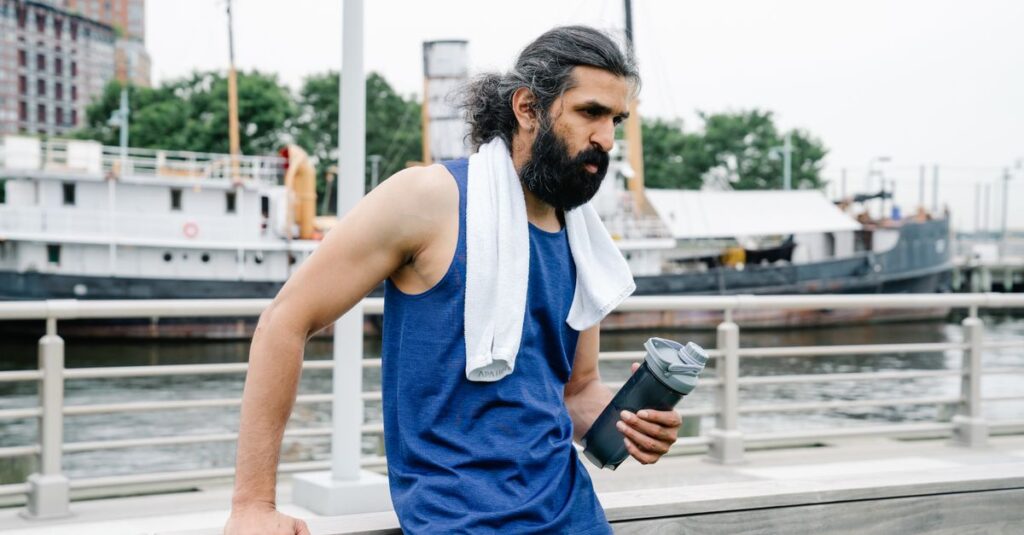 A man with a towel and water bottle rests by the waterfront after exercise.