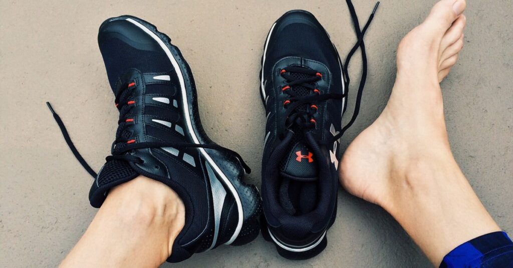 A pair of black sneakers next to bare feet on a gray surface.